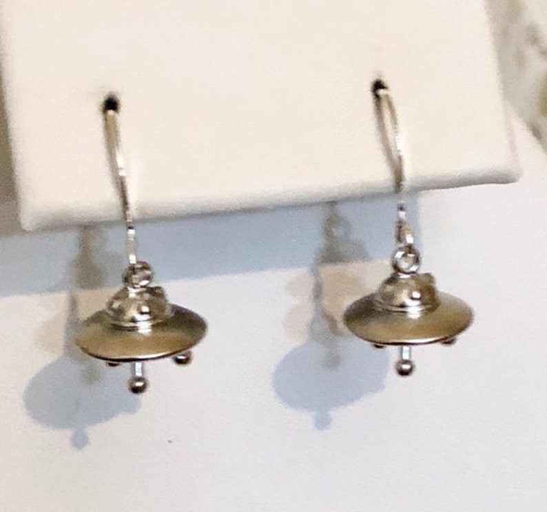 May include: A pair of silver earrings with a UFO design. The earrings are hanging from a silver hook.