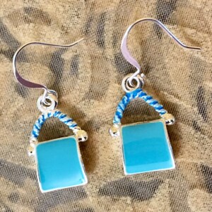 Pu&ograve; includere: Un paio di orecchini pendenti in argento con un ciondolo quadrato blu turchese e un dettaglio di corda blu e bianca.