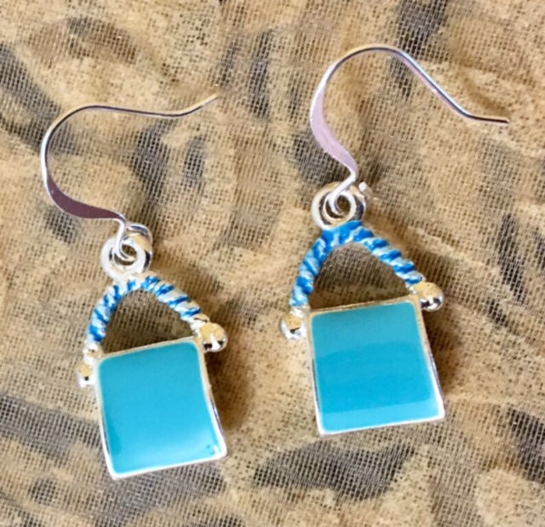 Pu&ograve; includere: Un paio di orecchini pendenti in argento con un ciondolo quadrato blu turchese e un dettaglio di corda blu e bianca.