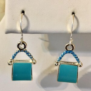Pu&ograve; includere: Un paio di orecchini pendenti in argento con un ciondolo quadrato blu turchese. Il ciondolo &egrave; circondato da un filo d'argento con un design intrecciato blu e bianco.