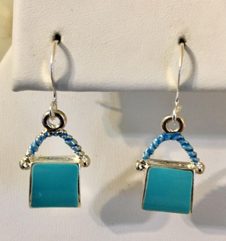 Pu&ograve; includere: Un paio di orecchini pendenti in argento con un ciondolo quadrato blu turchese. Il ciondolo &egrave; circondato da un filo d'argento con un design intrecciato blu e bianco.