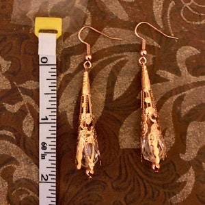 May include: A pair of rose gold filigree earrings with a teardrop shape. The earrings are approximately 2 inches long and feature a delicate, openwork design. Each earring has a clear, faceted crystal at the bottom, adding a touch of sparkle.