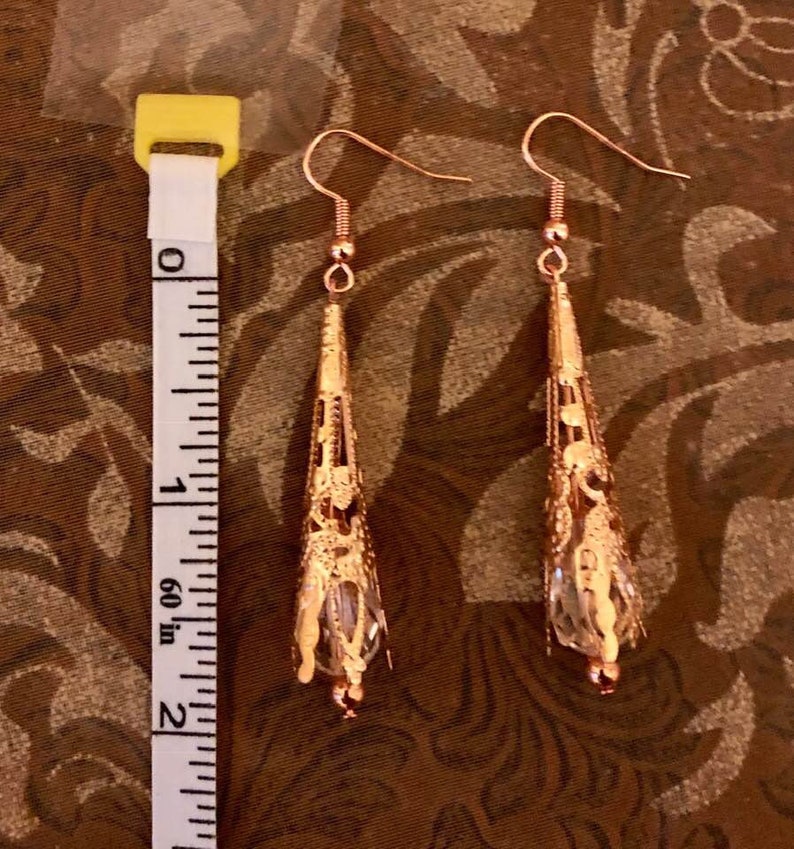 May include: A pair of rose gold filigree earrings with a teardrop shape. The earrings are approximately 2 inches long and feature a delicate, openwork design. Each earring has a clear, faceted crystal at the bottom, adding a touch of sparkle.