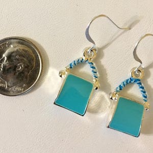 Pu&ograve; includere: Un paio di orecchini pendenti in argento con un ciondolo quadrato blu turchese. Il ciondolo &egrave; circondato da un design a corda blu e bianca.