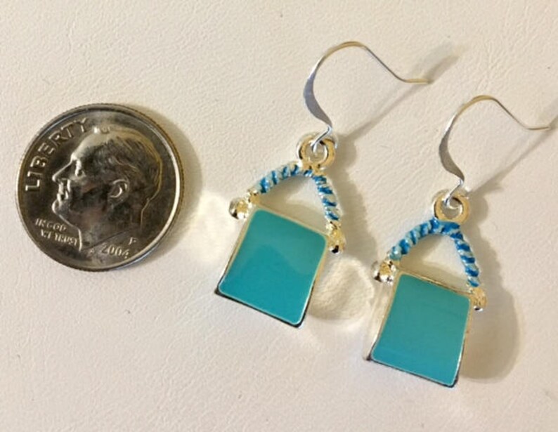 Pu&ograve; includere: Un paio di orecchini pendenti in argento con un ciondolo quadrato blu turchese. Il ciondolo &egrave; circondato da un design a corda blu e bianca.