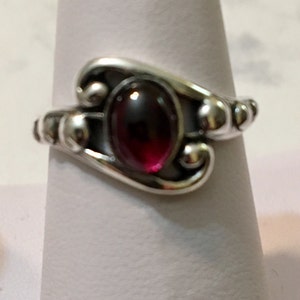 May include: A silver ring featuring a dark red gemstone set in a swirling design.