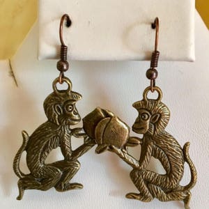 Puede incluir: Un par de pendientes de bronce con forma de mono con un diseño floral. Los monos sostienen la flor en sus manos.