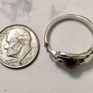 May include: A silver ring with a dark red gemstone set in a decorative design. The ring is next to a US dime for size comparison.