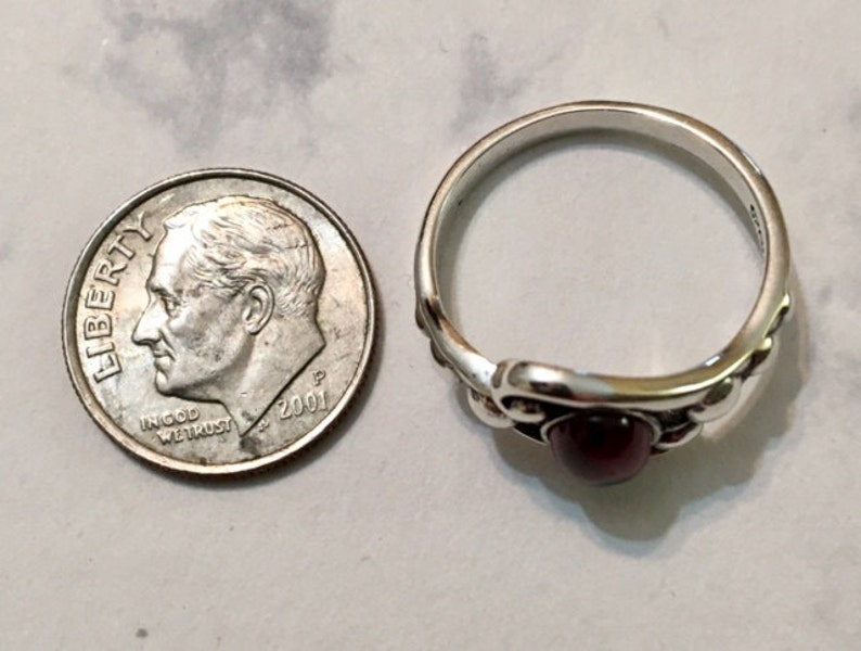 May include: A silver ring with a dark red gemstone set in a decorative design. The ring is next to a US dime for size comparison.
