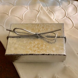 May include: A small, silver gift box with a white and gold patterned design. The box is tied with a silver ribbon and bow.