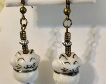 Puede incluir: Un par de pendientes colgantes con dijes de gato de porcelana blanca con detalles faciales negros. Los dijes están acentuados con cuentas doradas y plateadas y diamantes de imitación. Los pendientes tienen ganchos de bronce.