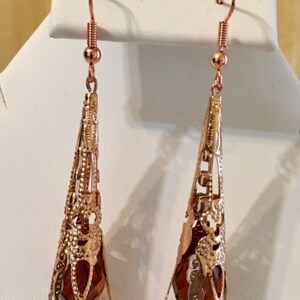 May include: A pair of copper-colored filigree earrings. Each earring features a cone-shaped design with intricate cut-out patterns and a faceted, teardrop-shaped amber-colored bead at the base. The earrings hang from a simple hook.