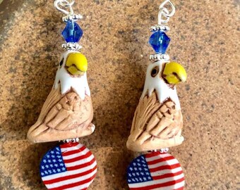 May include: Patriotic earrings featuring a ceramic bald eagle charm, a red, white, and blue American flag charm, and blue crystal beads.