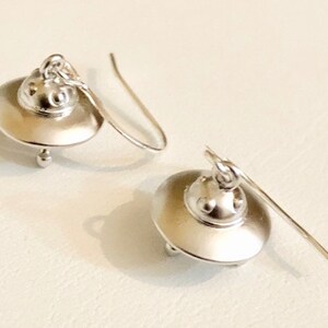 May include: A pair of silver UFO earrings. Each earring features a small, round UFO with a domed top and a small, round detail on the bottom. The earrings are attached to a silver hook.