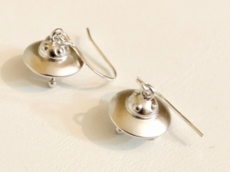 May include: A pair of silver UFO earrings. Each earring features a small, round UFO with a domed top and a small, round detail on the bottom. The earrings are attached to a silver hook.
