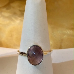Pu&ograve; includere: Un anello d'argento con una pietra ovale di colore viola chiaro. L'anello &egrave; su un espositore bianco.