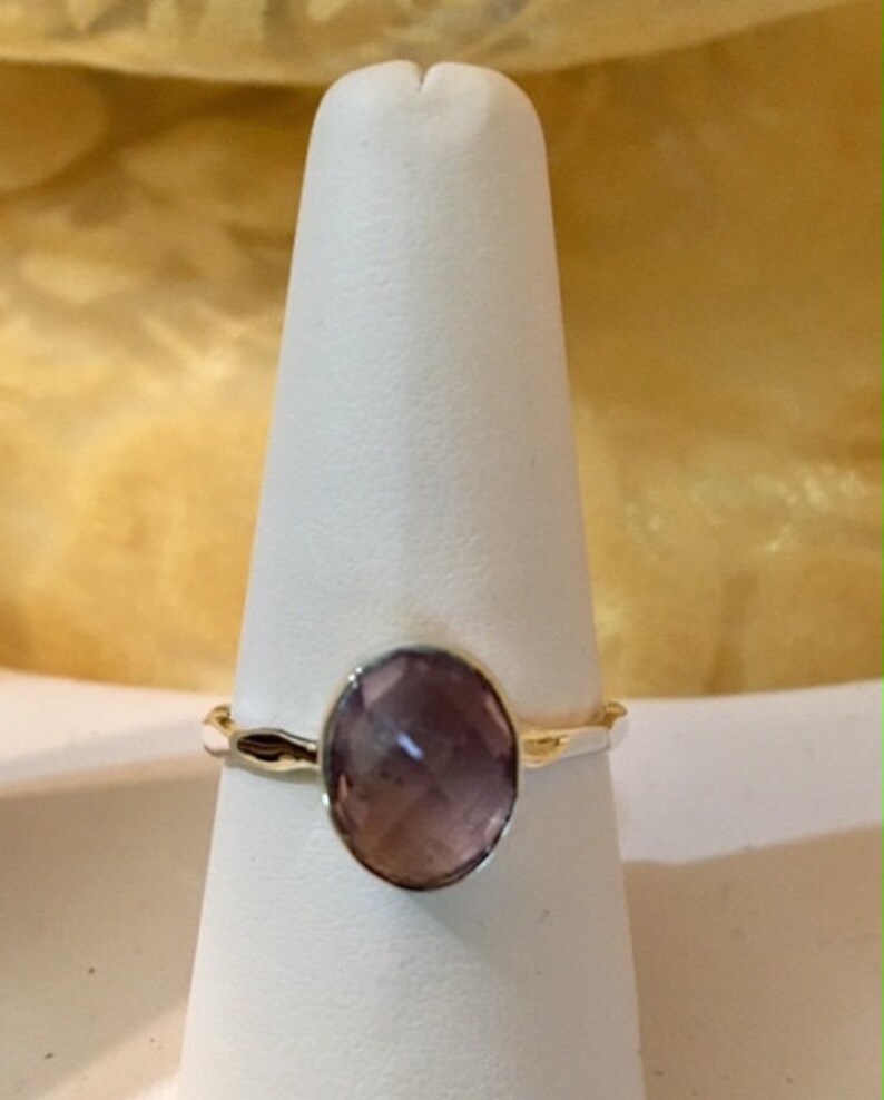 Pu&ograve; includere: Un anello d'argento con una pietra ovale di colore viola chiaro. L'anello &egrave; su un espositore bianco.