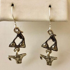 May include: Silver dangle earrings featuring a detailed charm of a bikini top and bottom.