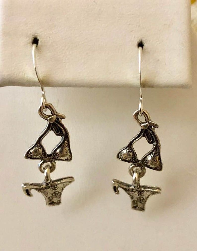 May include: Silver dangle earrings featuring a detailed charm of a bikini top and bottom.