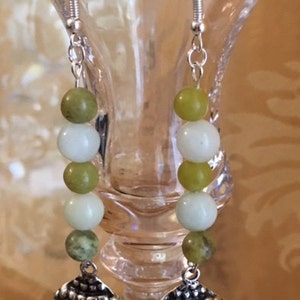 May include: Silver-toned dangle earrings featuring a Buddha head charm. Each earring has a series of white and green beads. The earrings are suspended from silver-colored ear wires.
