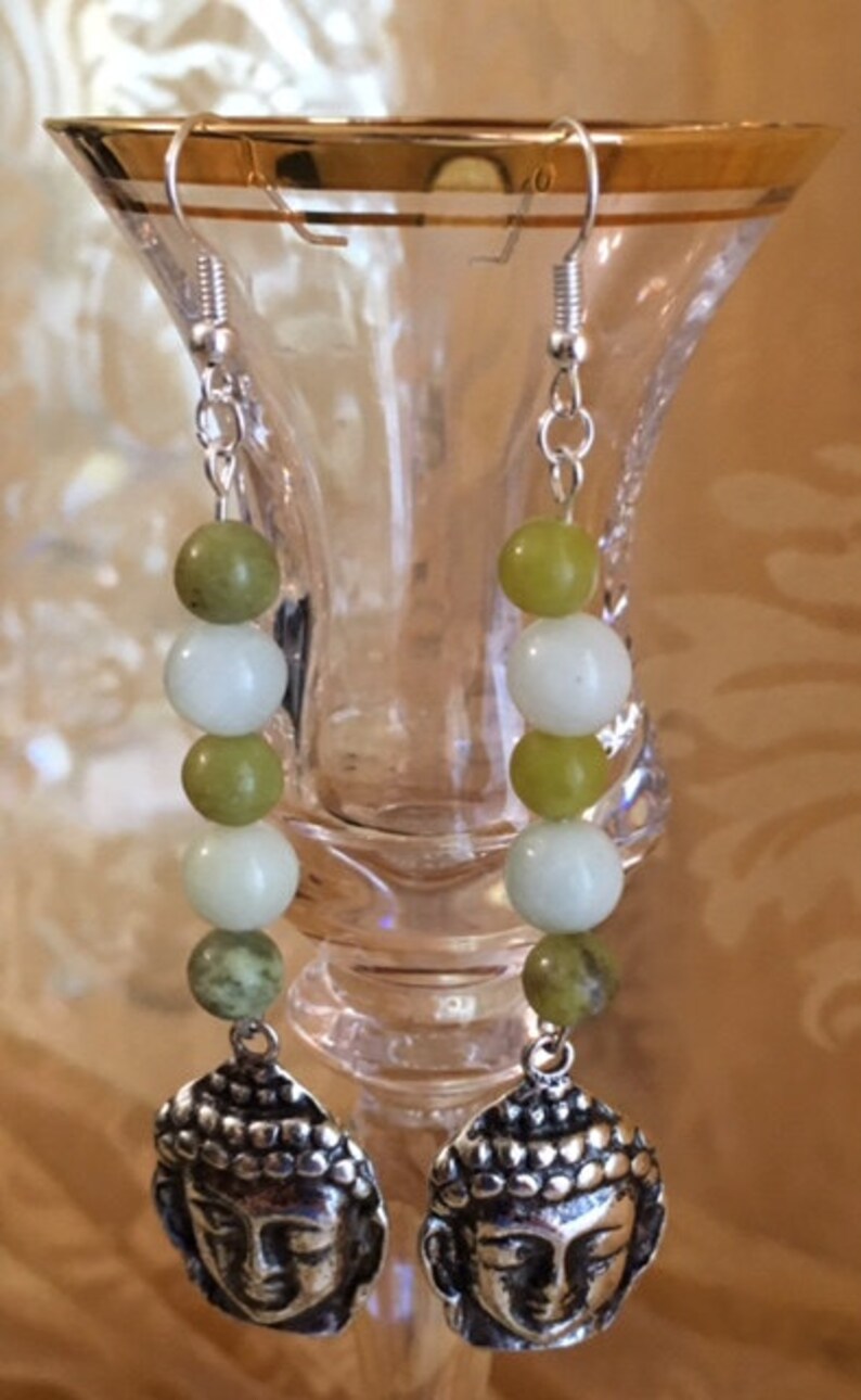 May include: Silver-toned dangle earrings featuring a Buddha head charm. Each earring has a series of white and green beads. The earrings are suspended from silver-colored ear wires.