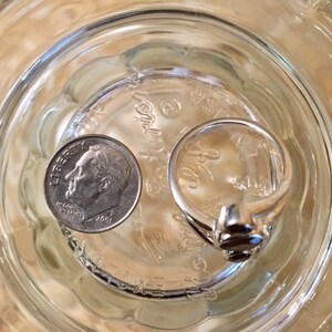 Peut inclure: Une bague en argent avec une pierre sombre sertie au centre. La bague est pos&eacute;e sur une surface en verre &agrave; c&ocirc;t&eacute; d'une pi&egrave;ce de dix cents am&eacute;ricaine dat&eacute;e de 2001.