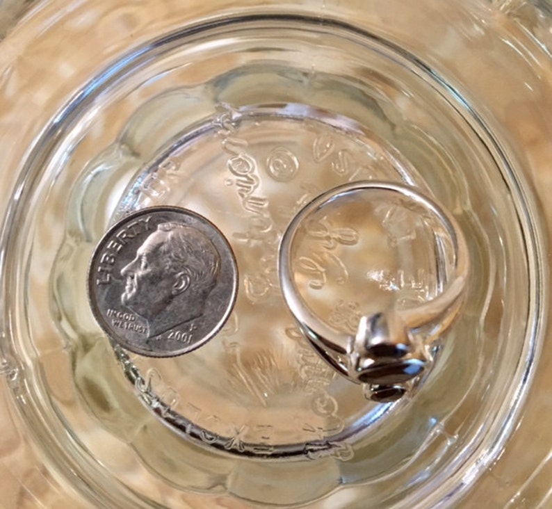 Peut inclure: Une bague en argent avec une pierre sombre sertie au centre. La bague est pos&eacute;e sur une surface en verre &agrave; c&ocirc;t&eacute; d'une pi&egrave;ce de dix cents am&eacute;ricaine dat&eacute;e de 2001.