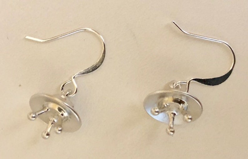 May include: A pair of silver earring findings with a small, round, flat base and three prongs. The findings have a hook closure.