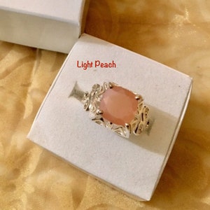 May include: A light peach colored ring with a detailed silver setting. The ring is displayed in a white box.