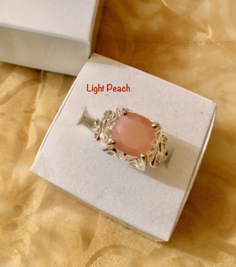 May include: A light peach colored ring with a detailed silver setting. The ring is displayed in a white box.