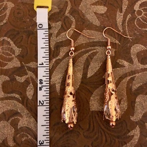 May include: A pair of copper-colored filigree earrings with a teardrop shape. The earrings are approximately 2.5 inches long, with a decorative pattern and a small bead at the bottom. The earrings hang from a simple hook.