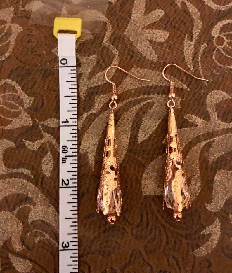May include: A pair of copper-colored filigree earrings with a teardrop shape. The earrings are approximately 2.5 inches long, with a decorative pattern and a small bead at the bottom. The earrings hang from a simple hook.