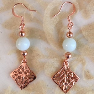 May include: A pair of rose gold earrings with a white bead and a diamond-shaped pendant with a filigree design.