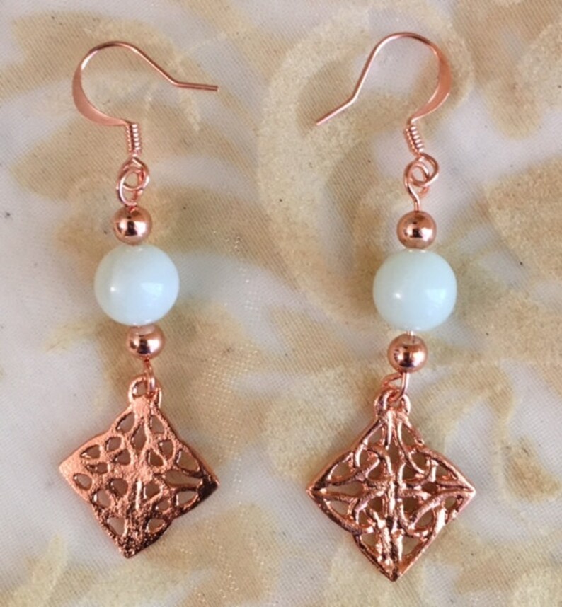 May include: A pair of rose gold earrings with a white bead and a diamond-shaped pendant with a filigree design.