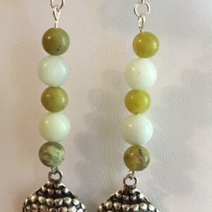 May include: A pair of dangle earrings featuring silver-toned Buddha head charms. Each earring has a series of green and white beads, with a silver hook for the ear. The earrings are designed for pierced ears.