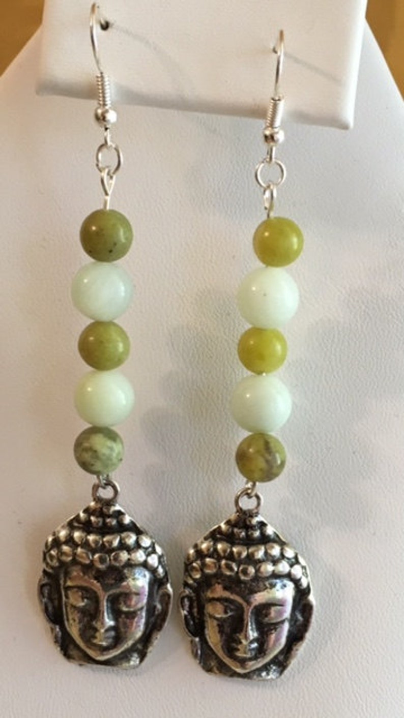 May include: A pair of dangle earrings featuring silver-toned Buddha head charms. Each earring has a series of green and white beads, with a silver hook for the ear. The earrings are designed for pierced ears.