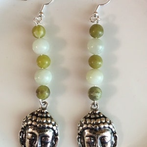 May include: A pair of dangle earrings featuring a silver Buddha head pendant. The earrings have a series of green and white beads strung together. The earrings are suspended from silver-colored ear hooks.