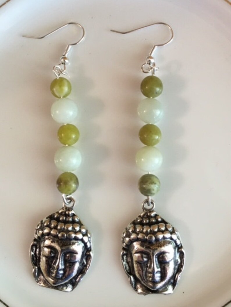 May include: A pair of dangle earrings featuring a silver Buddha head pendant. The earrings have a series of green and white beads strung together. The earrings are suspended from silver-colored ear hooks.