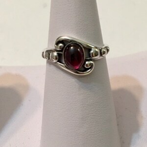 May include: A silver ring with a red gemstone set in a swirl design. The ring is on a white display stand.