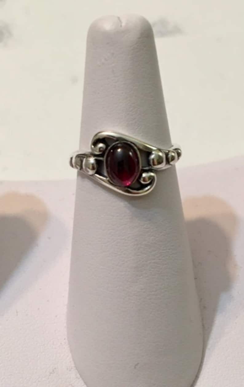 May include: A silver ring with a red gemstone set in a swirl design. The ring is on a white display stand.