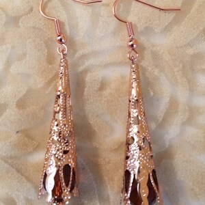 May include: A pair of rose gold filigree earrings. Each earring features a cone-shaped design with intricate cut-out patterns and a dark brown bead at the base. The earrings have a hook closure.