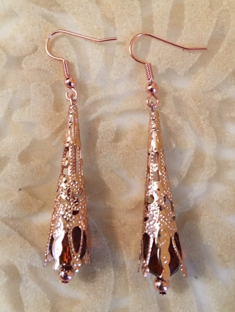 May include: A pair of rose gold filigree earrings. Each earring features a cone-shaped design with intricate cut-out patterns and a dark brown bead at the base. The earrings have a hook closure.