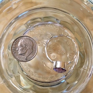 Pu&ograve; includere: Un anello d'argento con una pietra preziosa viola incastonata in una montatura a castone. L'anello &egrave; accanto a una moneta da dieci centesimi di dollaro americano.
