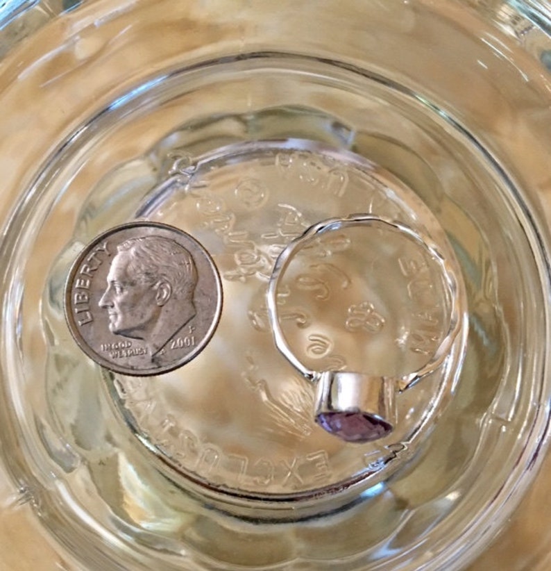 Pu&ograve; includere: Un anello d'argento con una pietra preziosa viola incastonata in una montatura a castone. L'anello &egrave; accanto a una moneta da dieci centesimi di dollaro americano.