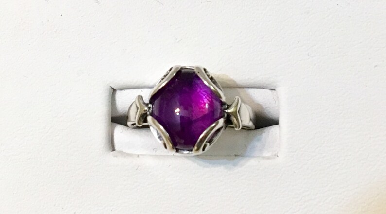 May include: A silver ring featuring a large, oval-shaped, deep purple gemstone. The gemstone is set in a decorative silver setting with ornate details. The ring is displayed on a white surface.