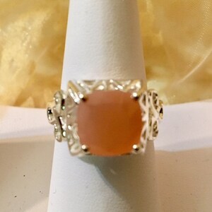 May include: A silver ring with a square, light pink gemstone set in a decorative, filigree design.