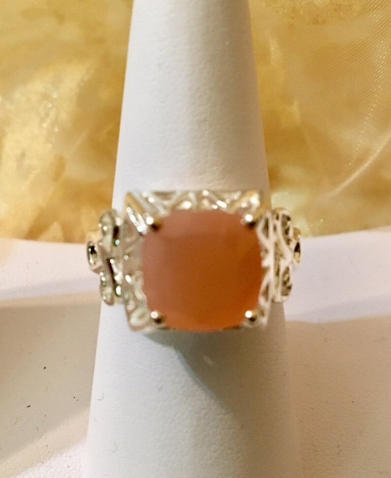 May include: A silver ring with a square, light pink gemstone set in a decorative, filigree design.