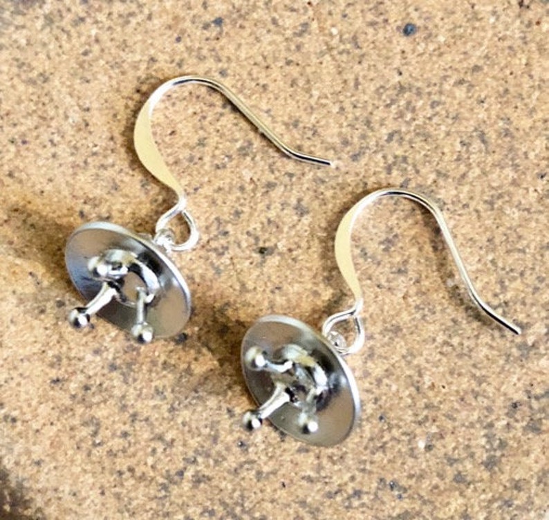 May include: A pair of silver earrings with a unique design. Each earring features a small, round disc with a raised, three-pronged design in the center.