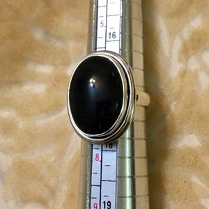Op de afbeelding: Een zilveren ring met een zwarte ovale edelsteen. De ring wordt op een ringmaat gelegd.