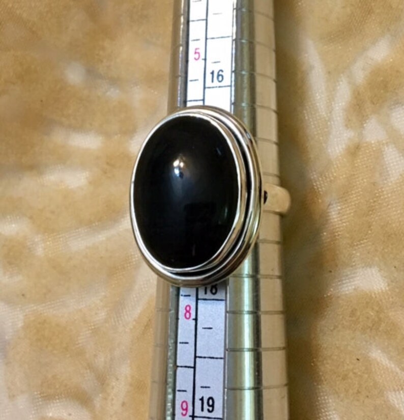 Op de afbeelding: Een zilveren ring met een zwarte ovale edelsteen. De ring wordt op een ringmaat gelegd.